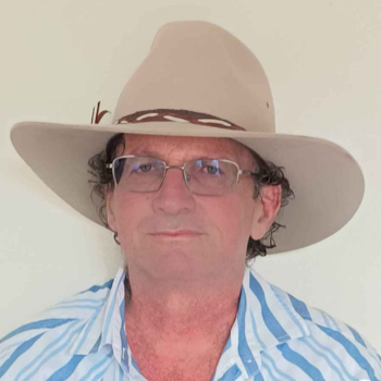 A man wearing a wide-brimmed hat and glasses, smiling in front of a plain background.